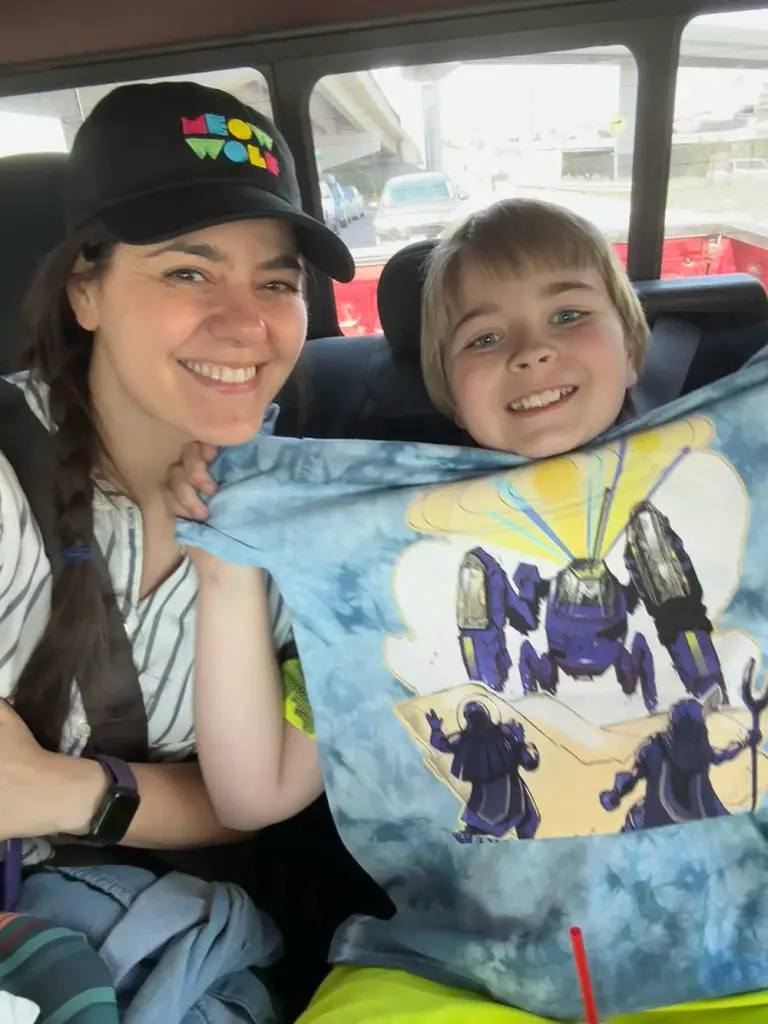 Photo of Stephanie in a black hat reading "Meow Wold" in bright colored letters and her nephew Maverick holding up a shirt with a robot from Meow Wolf pictured on it.