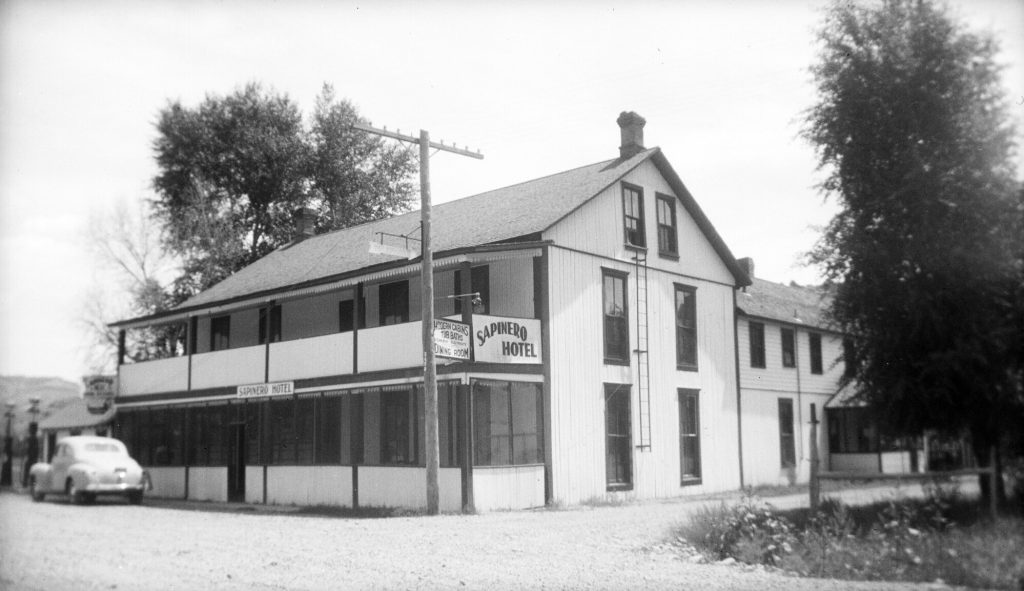 Photo of The Sapinero Hotel, in Sapinero (Gunnison County), Colorado, features a second story balcony, an open front porch, vertical board siding, intersecting rooflines, and fire escape ladder on the near wall. An attached sign reads: "Modern Cabins, Tub Baths, Electricity, Dining Room."