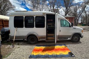 Photo of a shuttle van converted into a camper van with its door open and a bright and colorful RV mat outside for blog titled Exploring the World of Work Camping: Embracing Adventure and Flexibility.