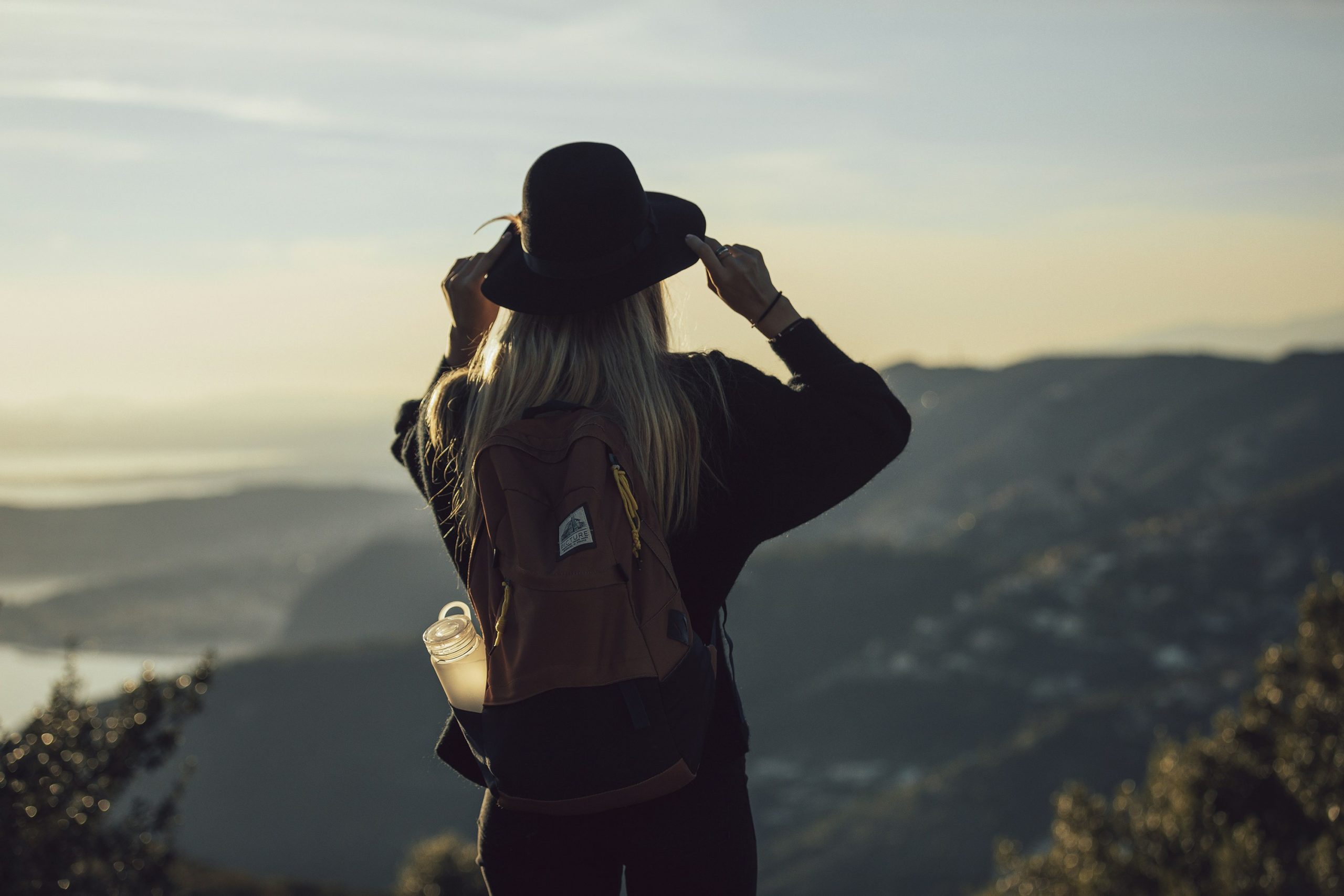 Featured image graphic for Applied Worldwide blog titled "Solo Travel Tips For The Fearless Wanderer" showing a female traveler looking out from the top of a mountain with a hat and backpack.