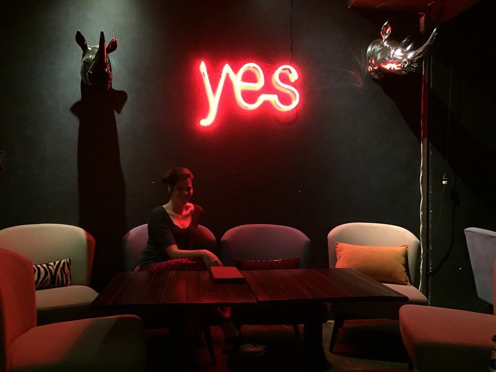 Photo of Stephanie sitting on a couch in a bar in Ho Chi Min City that is dimly lit by a red neon sign reading YES