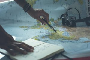 Photo of a a journal, a hand holding a pen, and a camera all on top of a large world map for Applied Worldwide blog "The Art of Maximizing Your Travel Adventures"